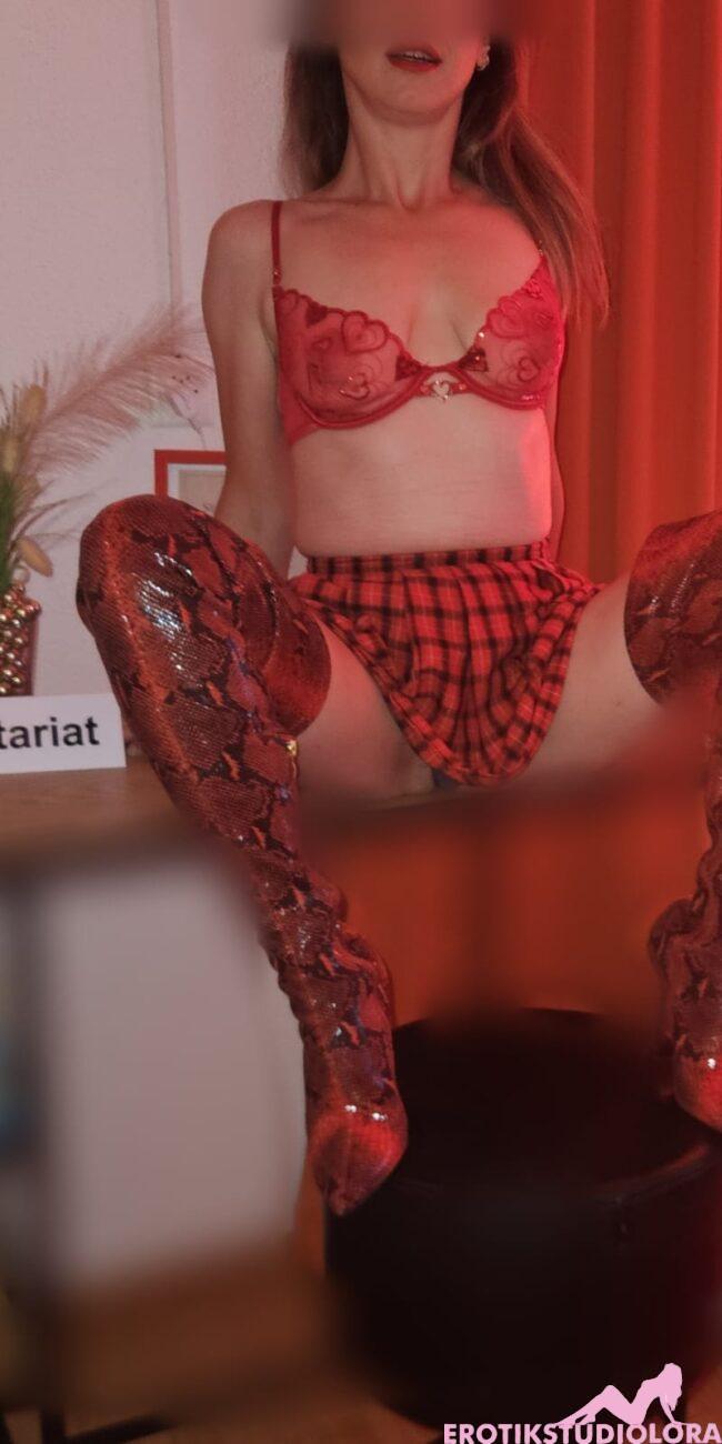 Woman in red lace bra and plaid skirt sits on a stool with legs apart, red curtains behind, and a 'Sekretariat' sign on the desk.